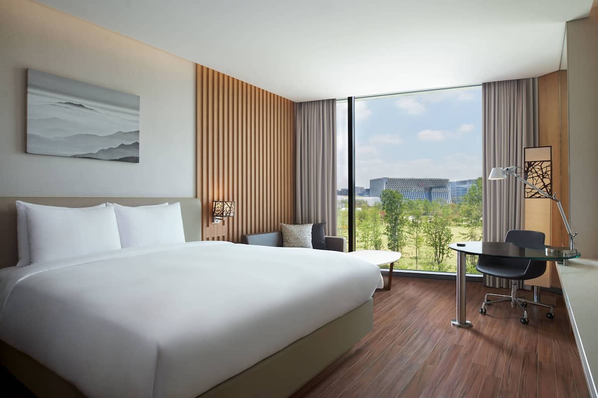 Ein modernes Hotelzimmer mit einem großen Bett, einem Schreibtisch mit Stuhl und Blick auf die Stadt durch große Fenster.