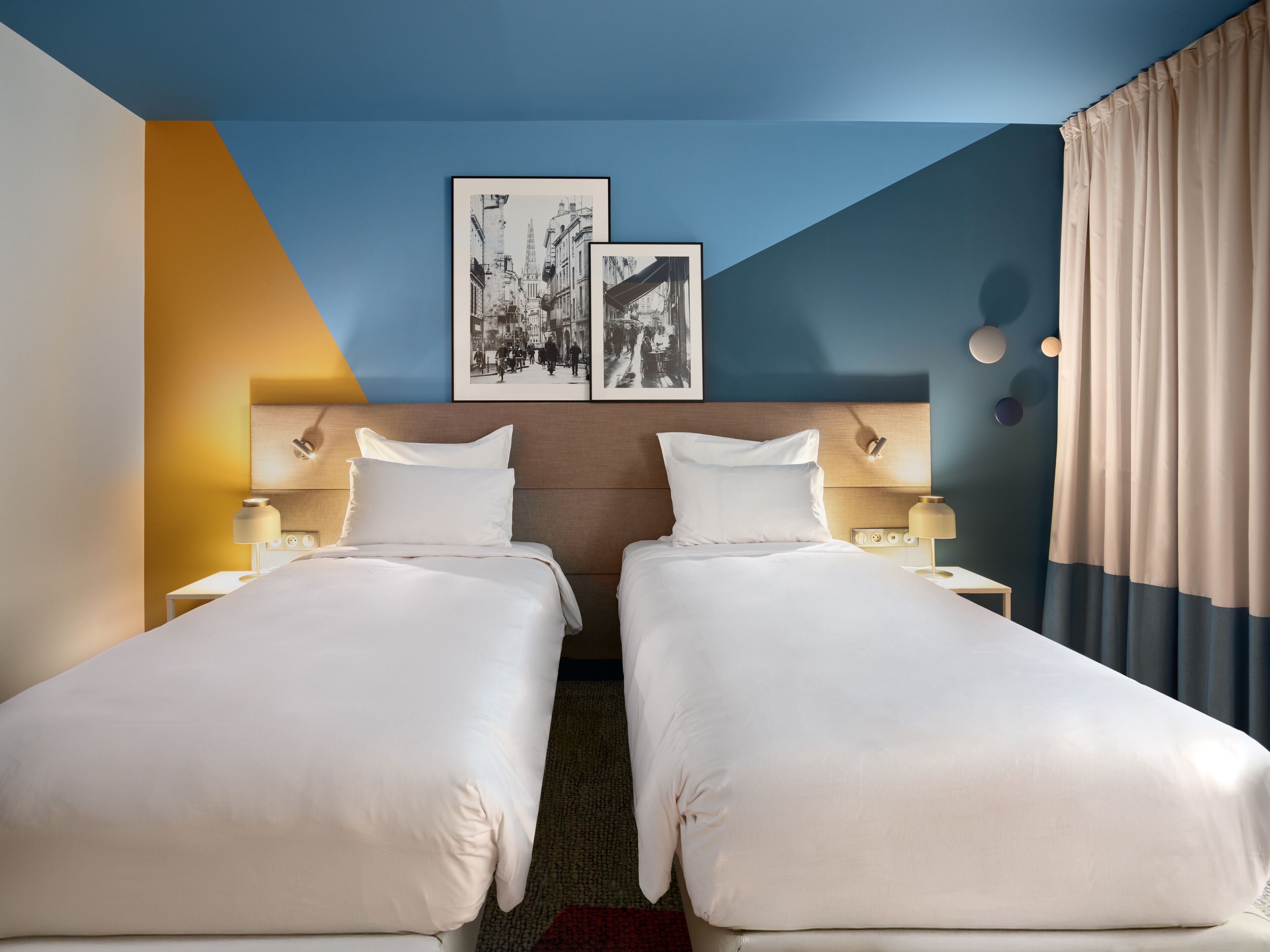 A hotel room with two beds, a headboard with a cityscape photo, bedside lamps, and a large window with curtains.