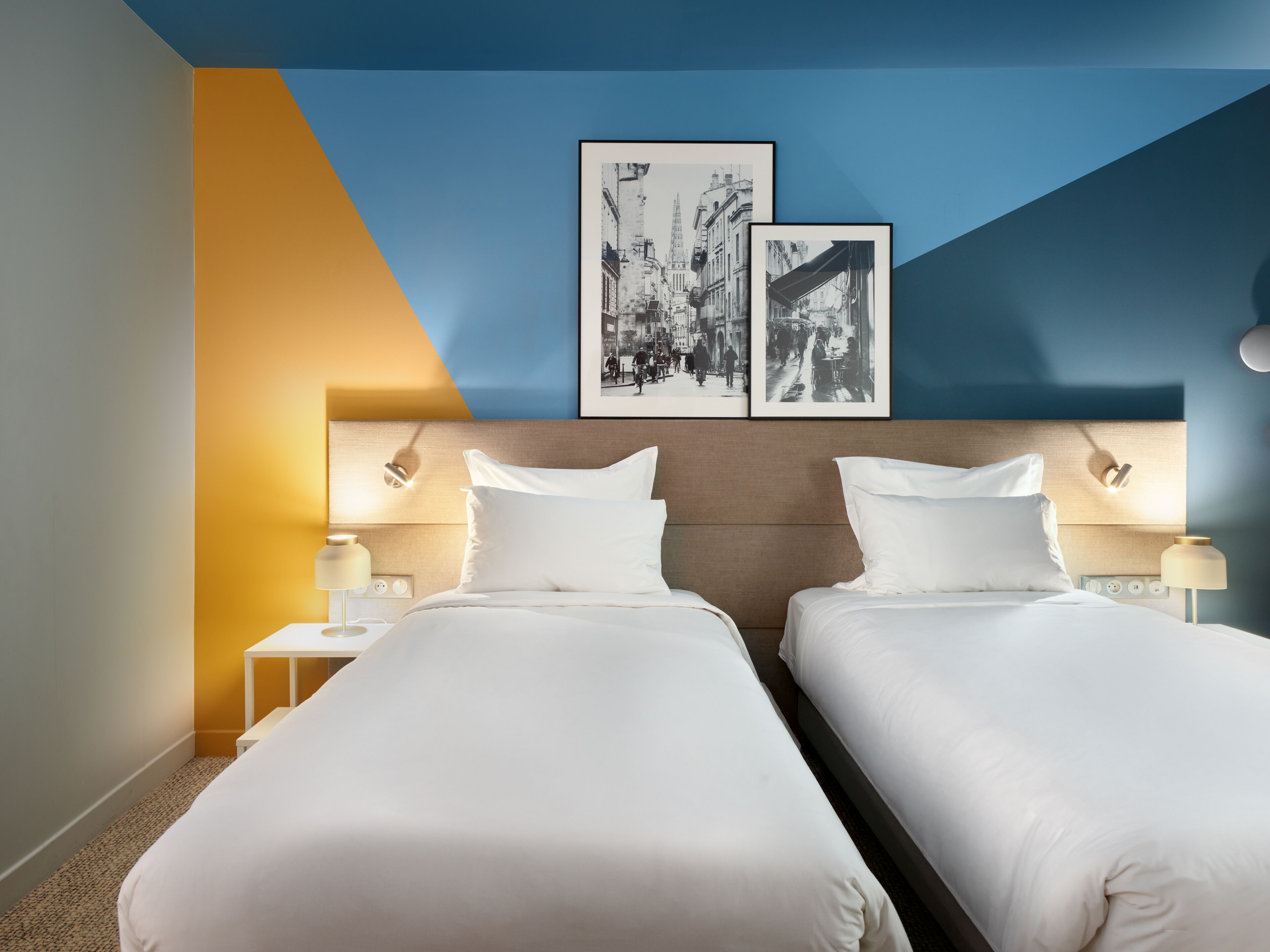 A hotel room with two beds, a headboard, bedside lamps, and two framed black and white photographs on the wall.