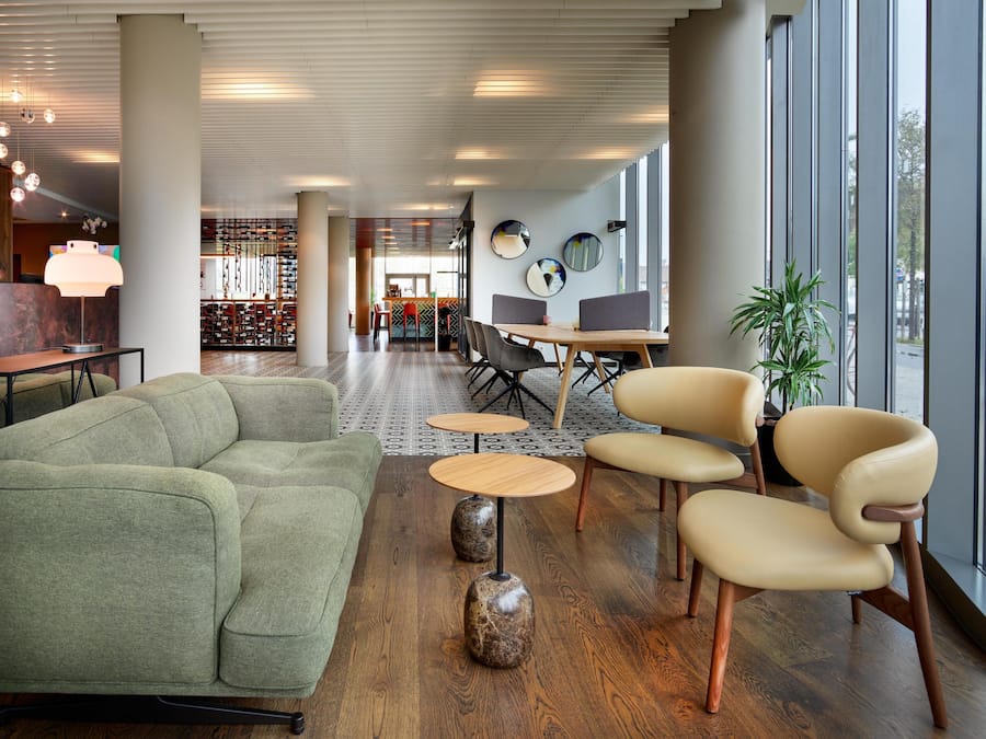 Un área de descanso moderna con un sofá verde, mesas redondas de madera y una mesa de comedor con sillas.