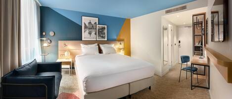 A modern hotel room with a large bed, a desk, and a chair. There is a window with curtains, a wall-mounted shelf, and a door leading to another room.