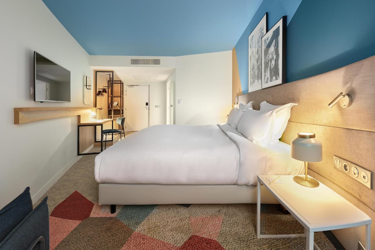A modern hotel room with a large bed, bedside table with a lamp, a TV mounted on the wall, and a desk with a chair.