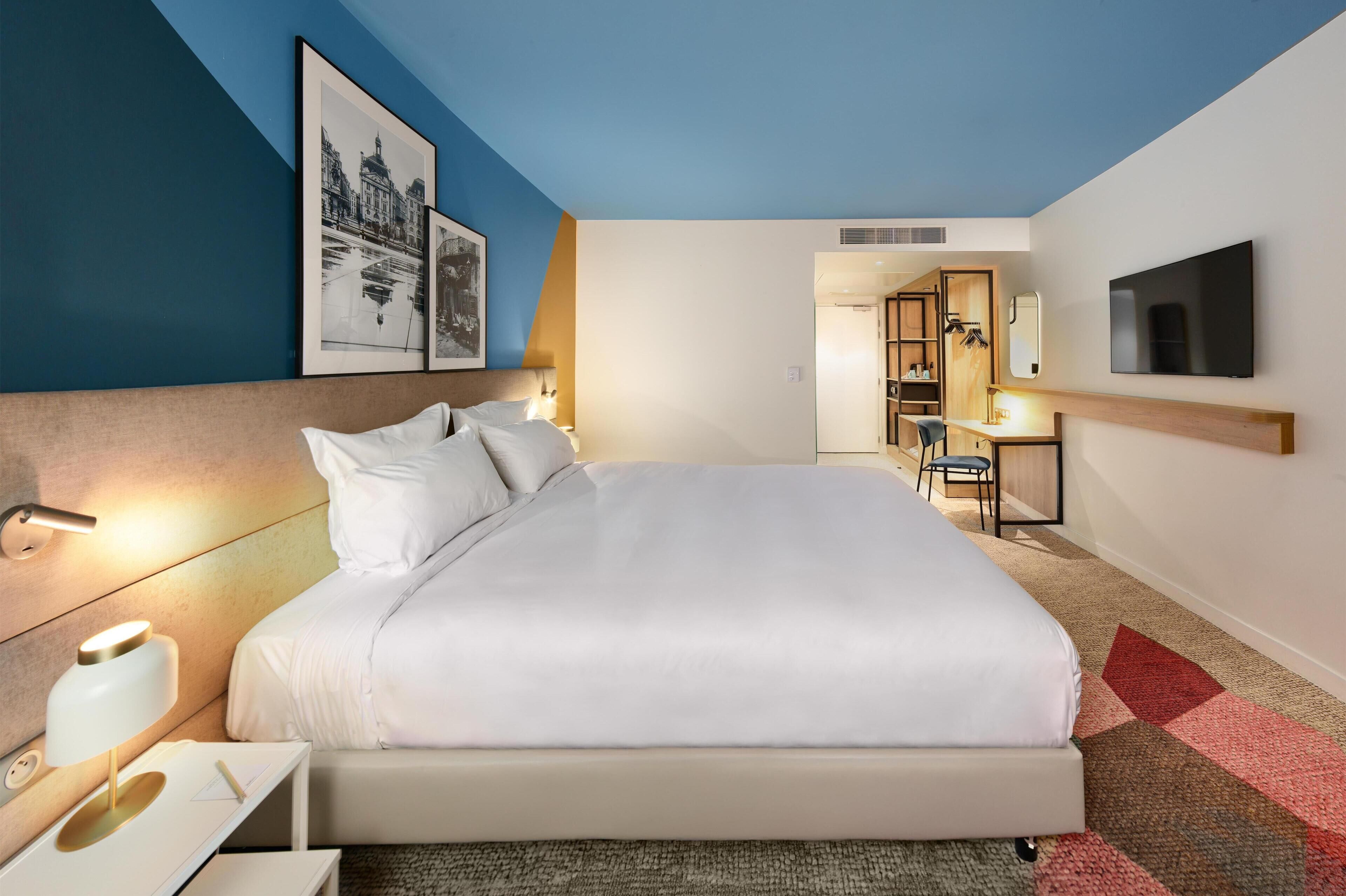 A modern hotel room with a large bed, bedside tables, a desk, and a TV.