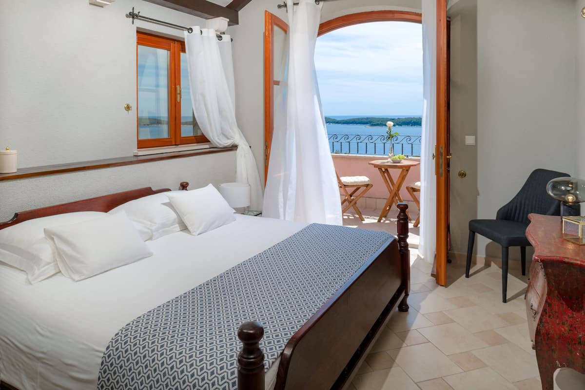 A bedroom with a large bed, a wooden headboard, a window with curtains, and a view of the sea.