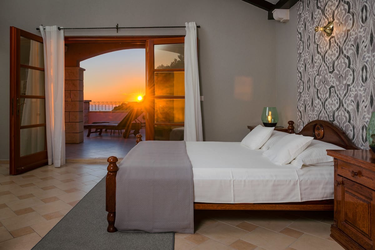 A bedroom with a bed, a nightstand, a lamp, and a view of the sunset through an open door.