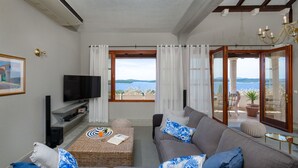 A living room with a sofa, a television, a wicker coffee table with a book and a glass of orange juice, and a view of the sea through a large window. - Hvar Boutique Villas (Hvar)