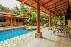 Un área de piscina con baldosas de piedra, un porche cubierto con sillas y una pérgola de madera.