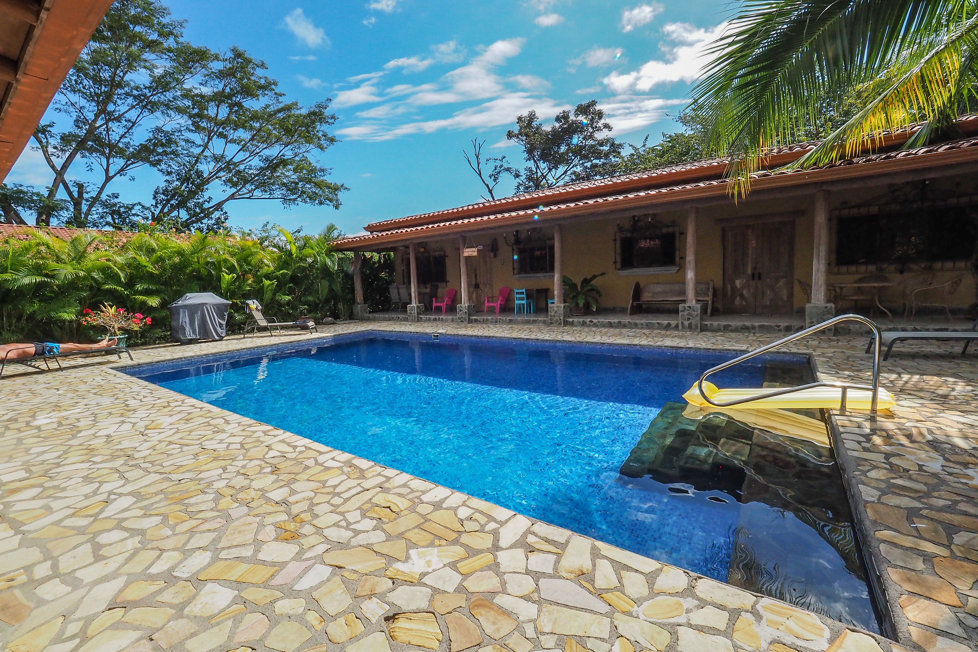 A swimming pool with stone tiles, a covered patio with chairs, and a building with a tiled roof.