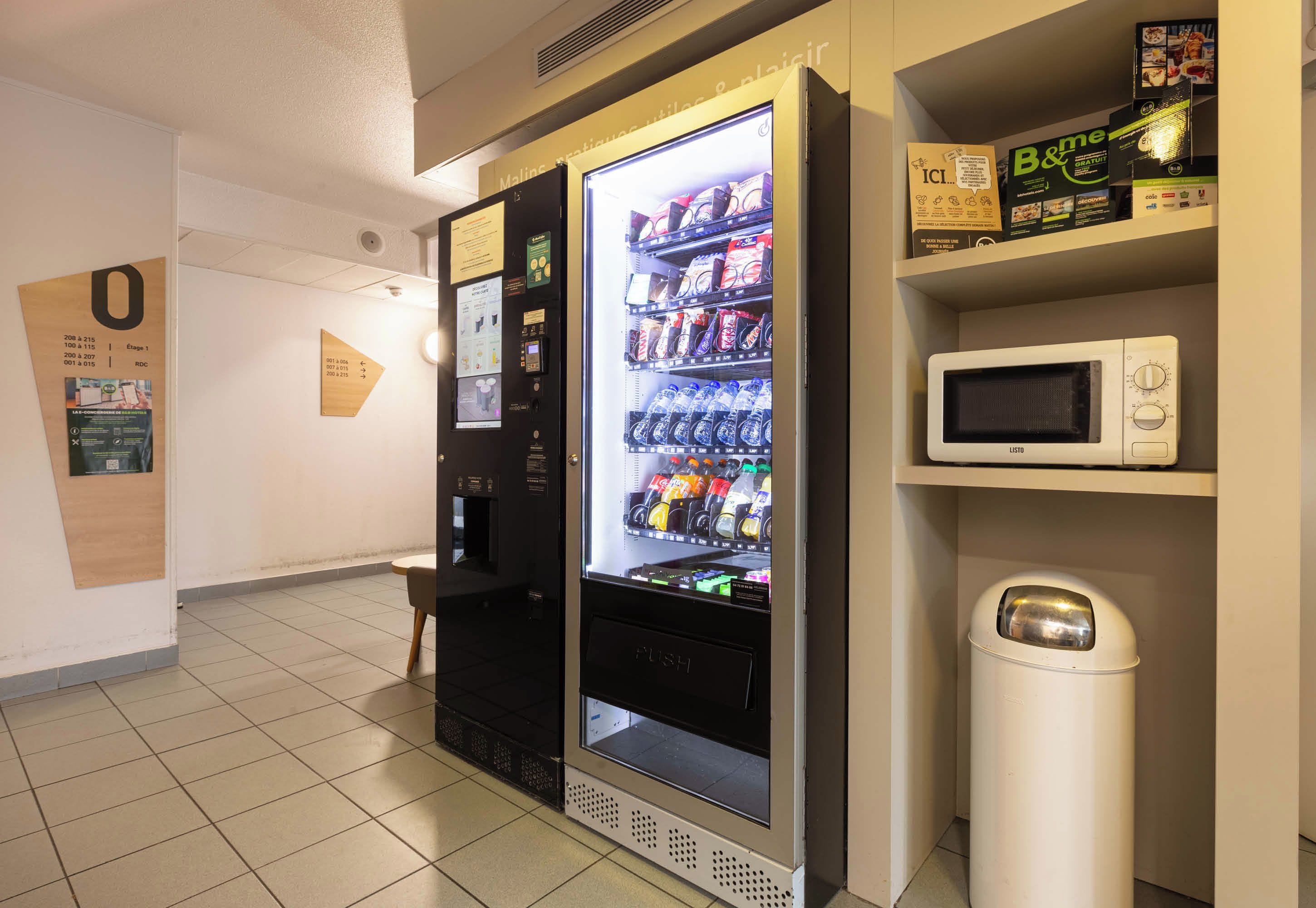 Máquina expendedora con opciones de bebidas, un microondas y un cubo de basura en un pasillo.