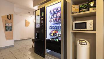 Máquina expendedora con opciones de bebidas, un microondas y un cubo de basura en un pasillo.