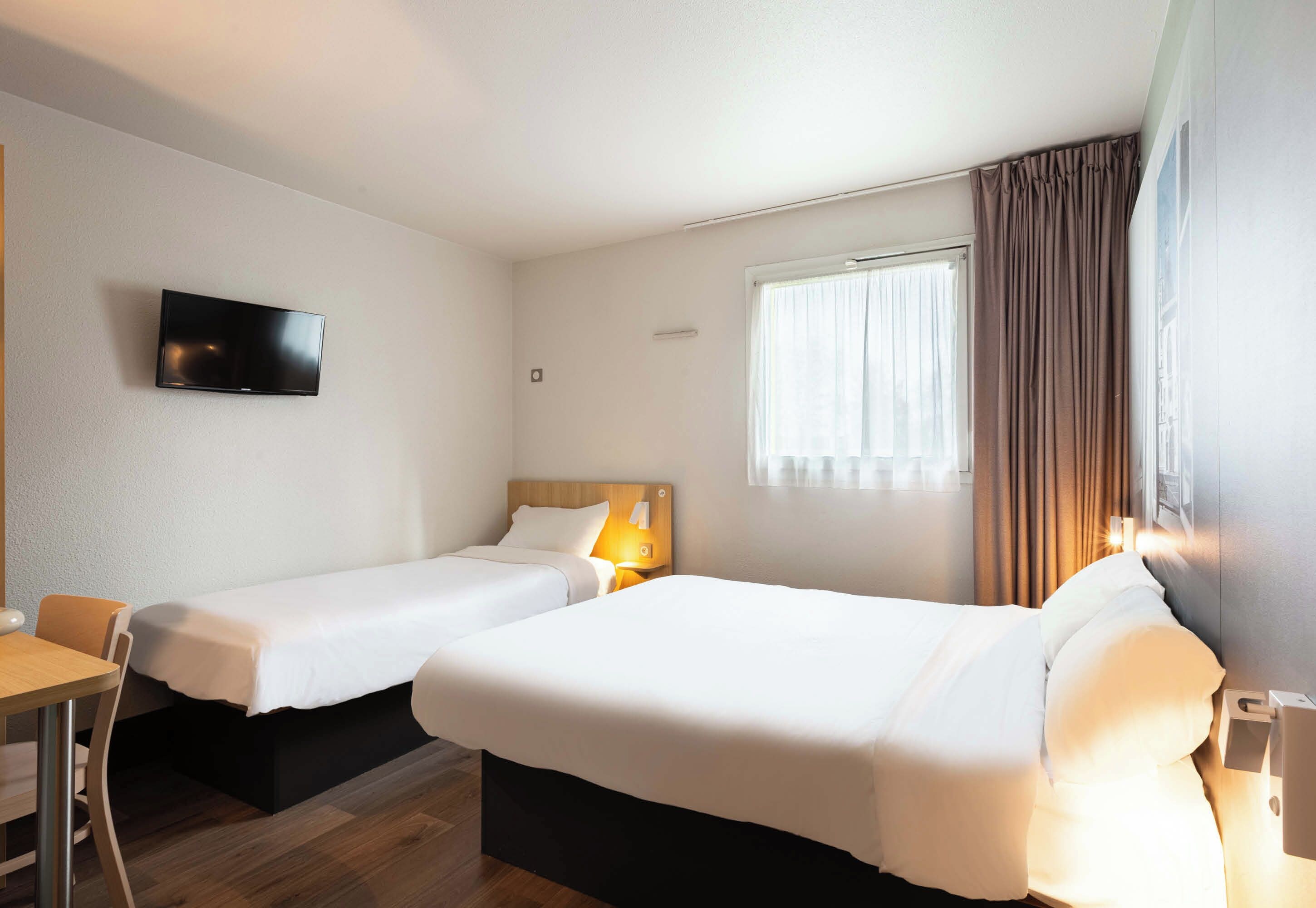 A hotel room with two single beds, a TV mounted on the wall, a desk, and a window with curtains.