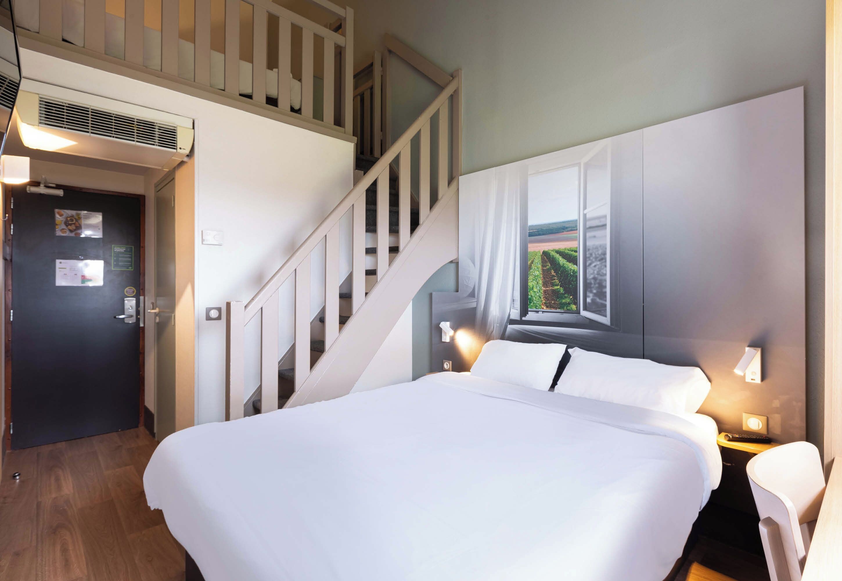 A hotel room with a large bed, a staircase, a window with a view, and a wall-mounted light.