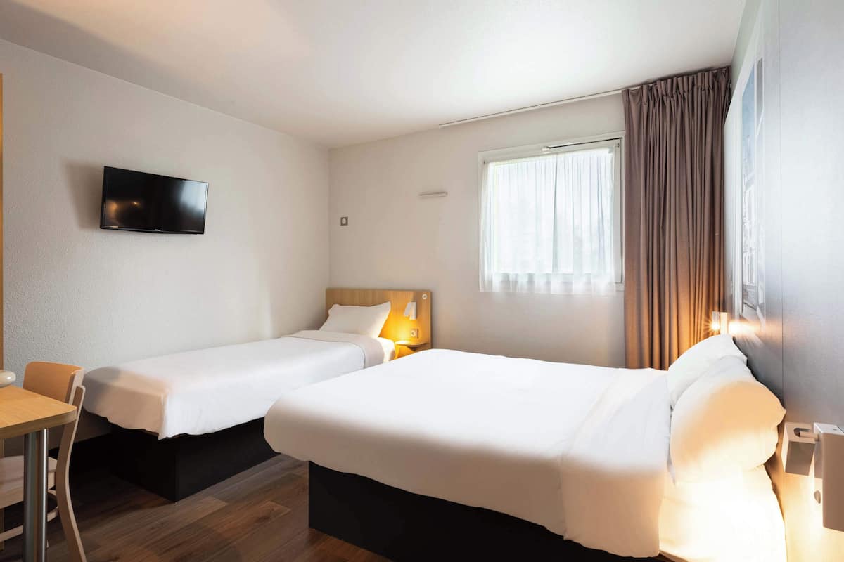 Ein Hotelzimmer mit zwei Einzelbetten, einem an der Wand montierten Fernseher, einem Schreibtisch und einem Fenster mit Vorhängen.