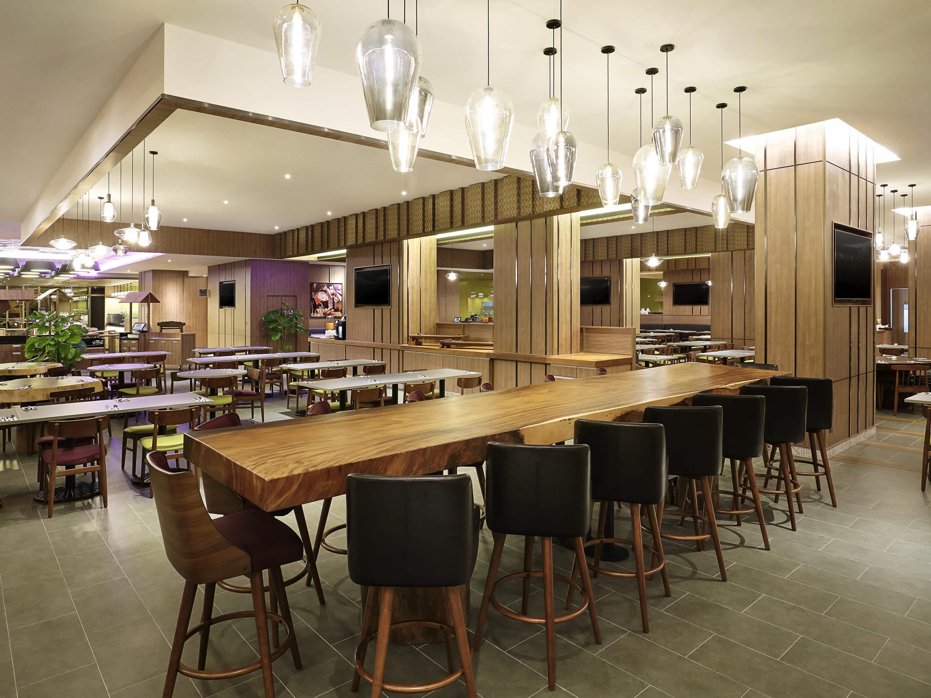 Un intérieur de restaurant moderne, avec un long bar en bois, plusieurs espaces de détente et des luminaires suspendus.