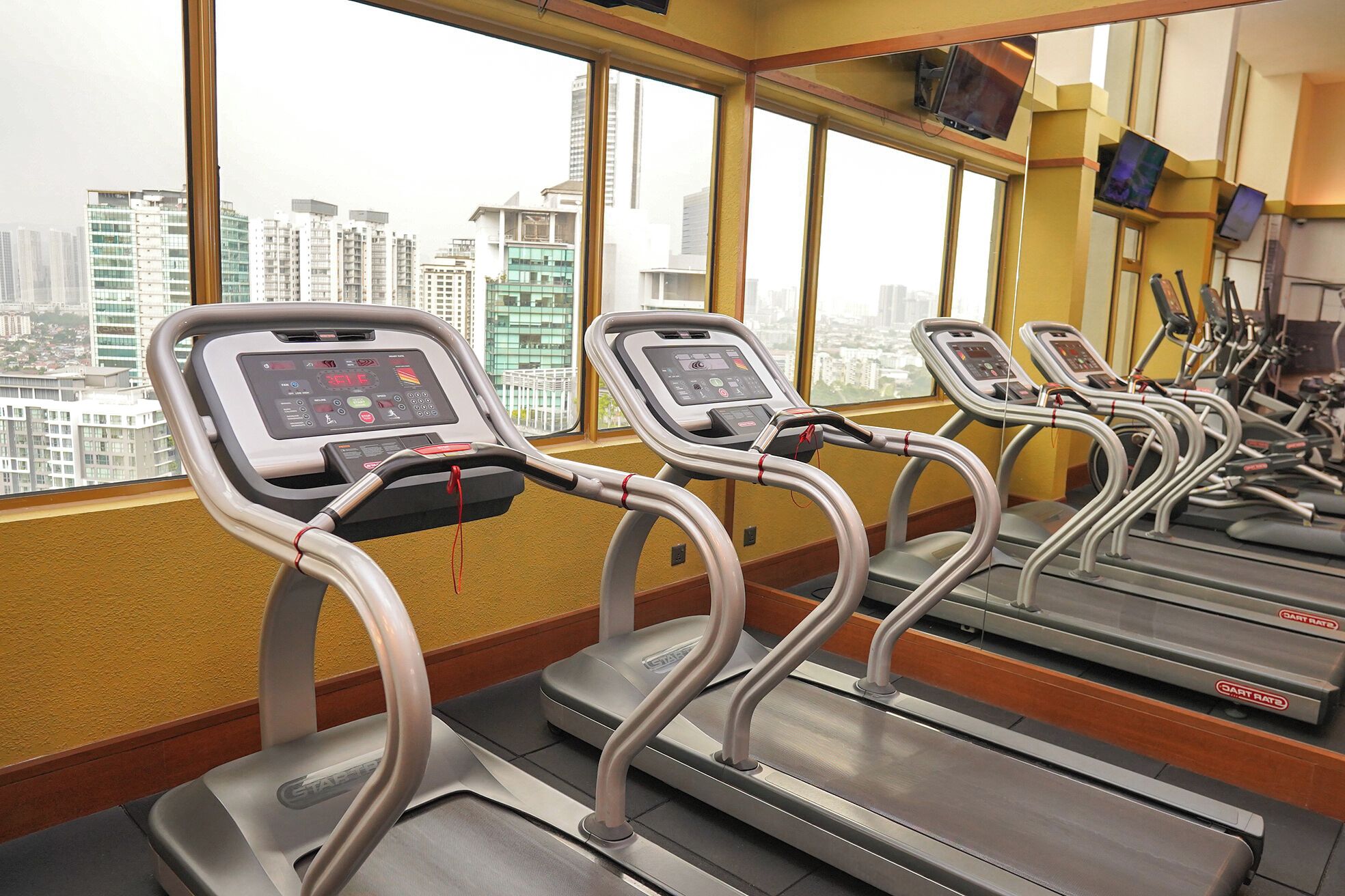 Un gimnasio con cintas de correr y equipo de ejercicio, amplias ventanas con vista a la ciudad y una pared amarilla.
