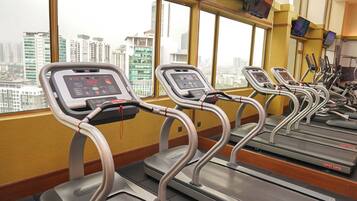 Un gimnasio con cintas de correr y equipo de ejercicio, amplias ventanas con vista a la ciudad y una pared amarilla.