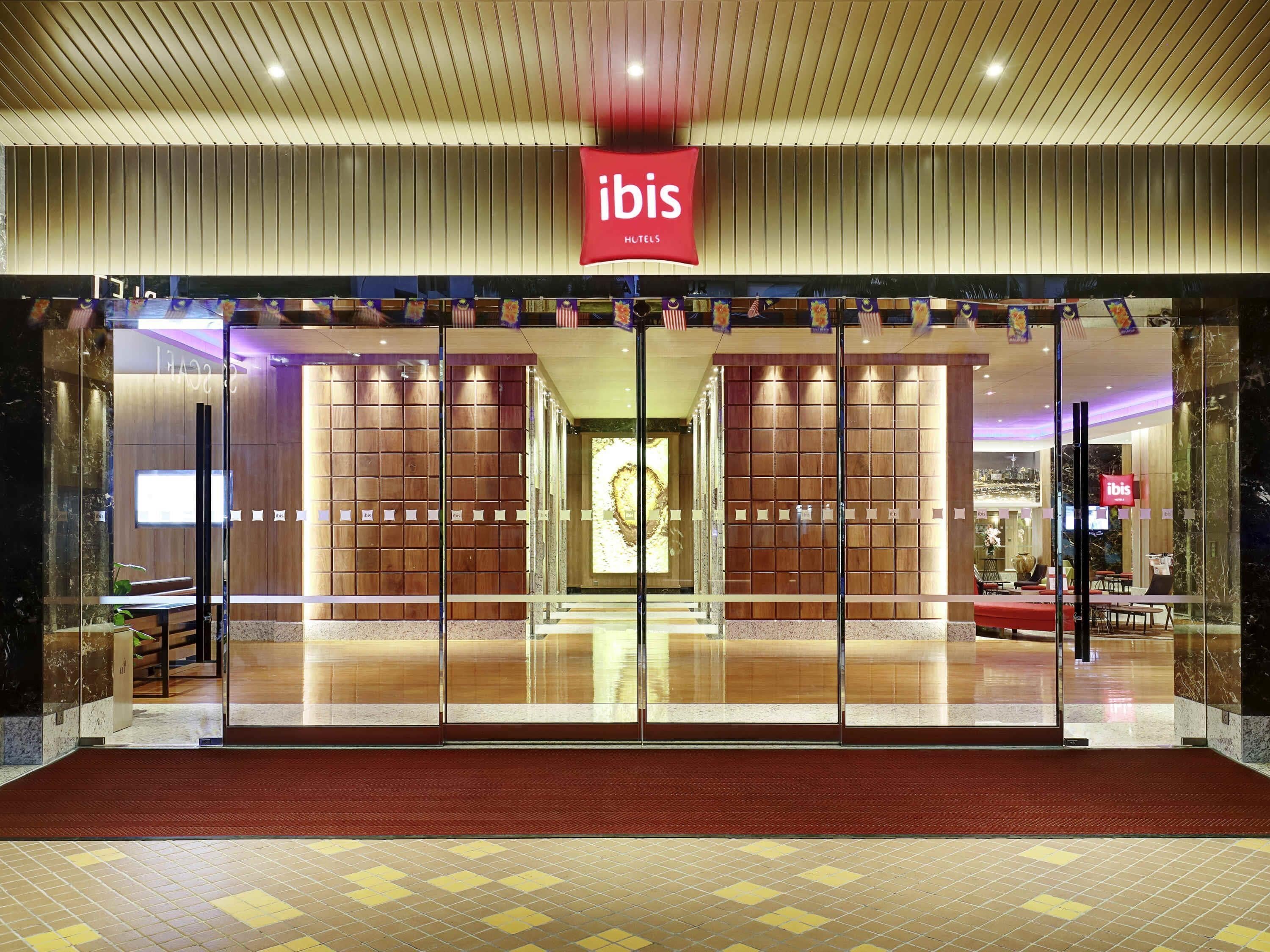 Entrada de hotel con un letrero rojo de 'ibis', puertas de vidrio y una alfombra roja que lleva al interior.