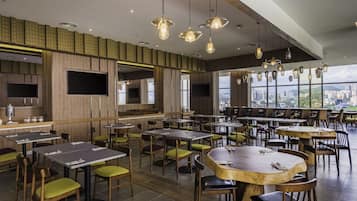 A modern restaurant with multiple dining tables and chairs, large windows offering city views, and a variety of hanging pendant lights.