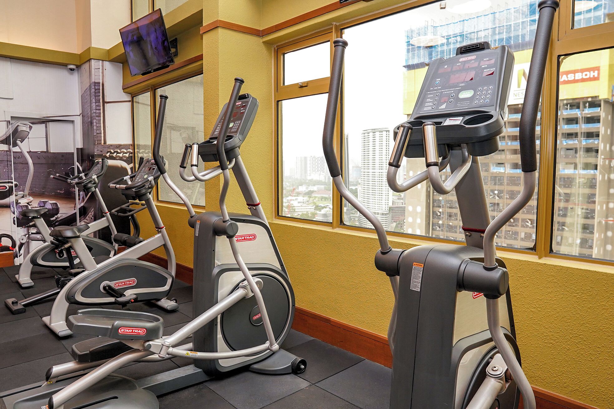 Una palestra con diverse macchine ellittiche StarTrac, un tapis roulant e una finestra vista città.