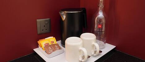 A coffee station with instant coffee packets, two white mugs, a spoon, a black electric kettle, and a glass bottle labeled 'ibis' on a countertop.