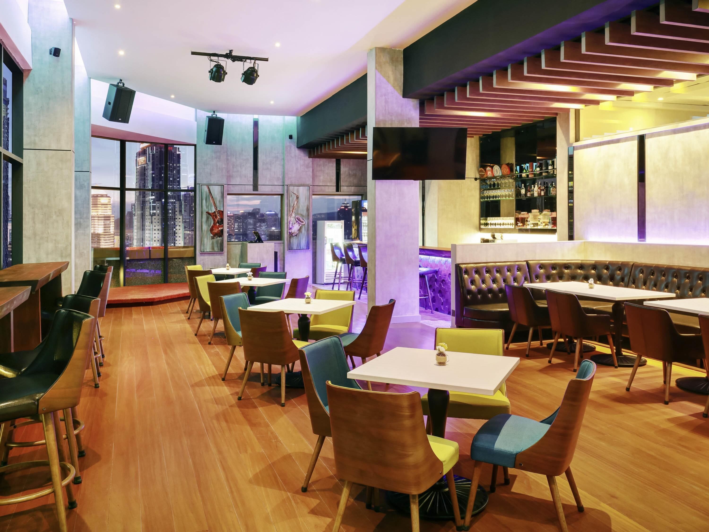 Um restaurante moderno com área de bar, diversas opções de assento incluindo mesas e cabines, e amplas janelas com vista para a cidade.