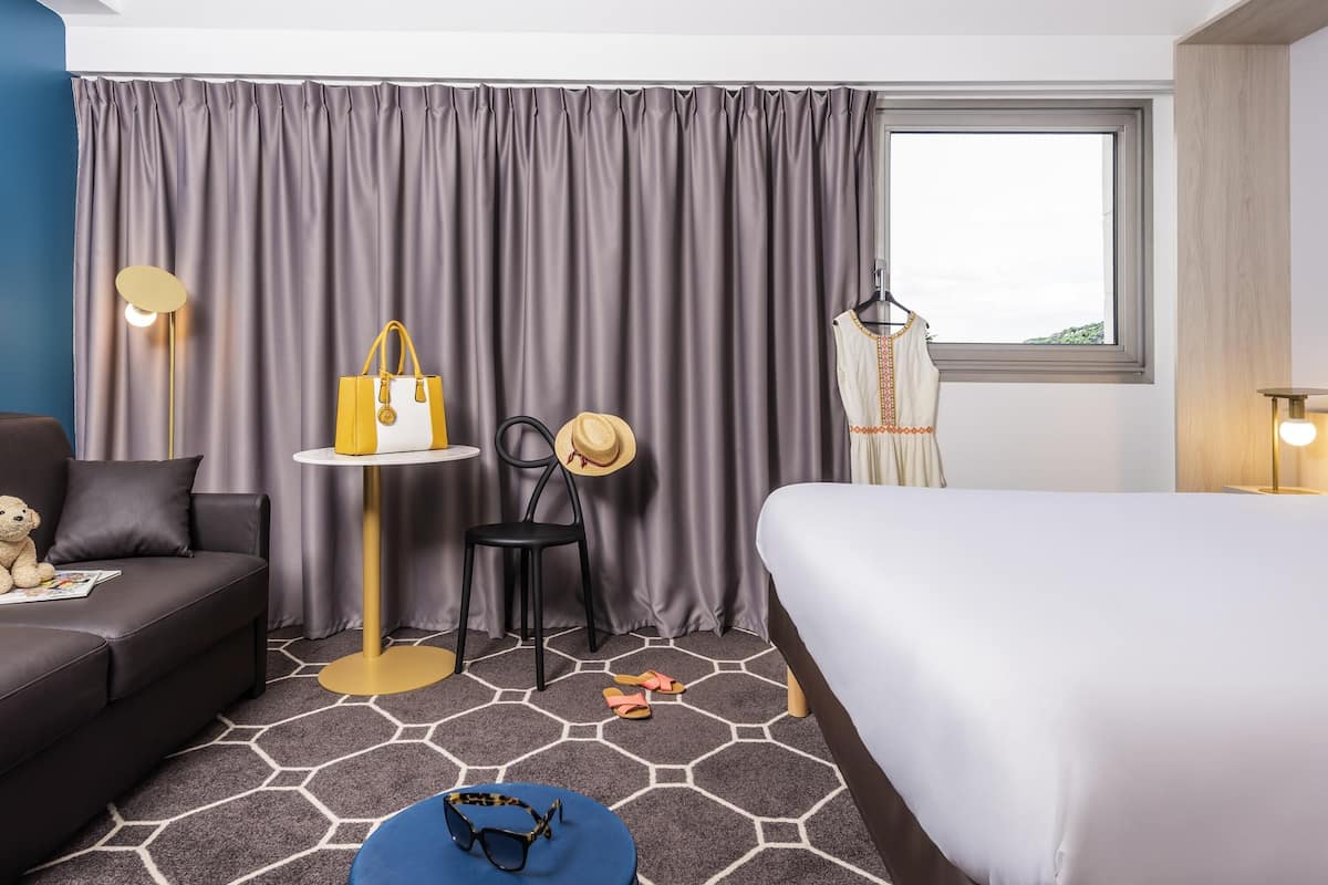 A hotel room with a bed, a sofa, a small table, a chair, a lamp, a hat, a handbag, and a teddy bear.