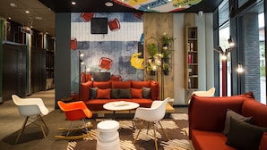 A modern lounge area with a red sofa, white chairs, and a small table. There are plants and bookshelves in the background. - ibis Vilnius Centre (Vilnius)