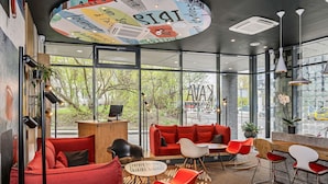 A modern interior with a red sofa, a wooden desk, and a large window offering a view of the street. - ibis Vilnius Centre (Vilnius)
