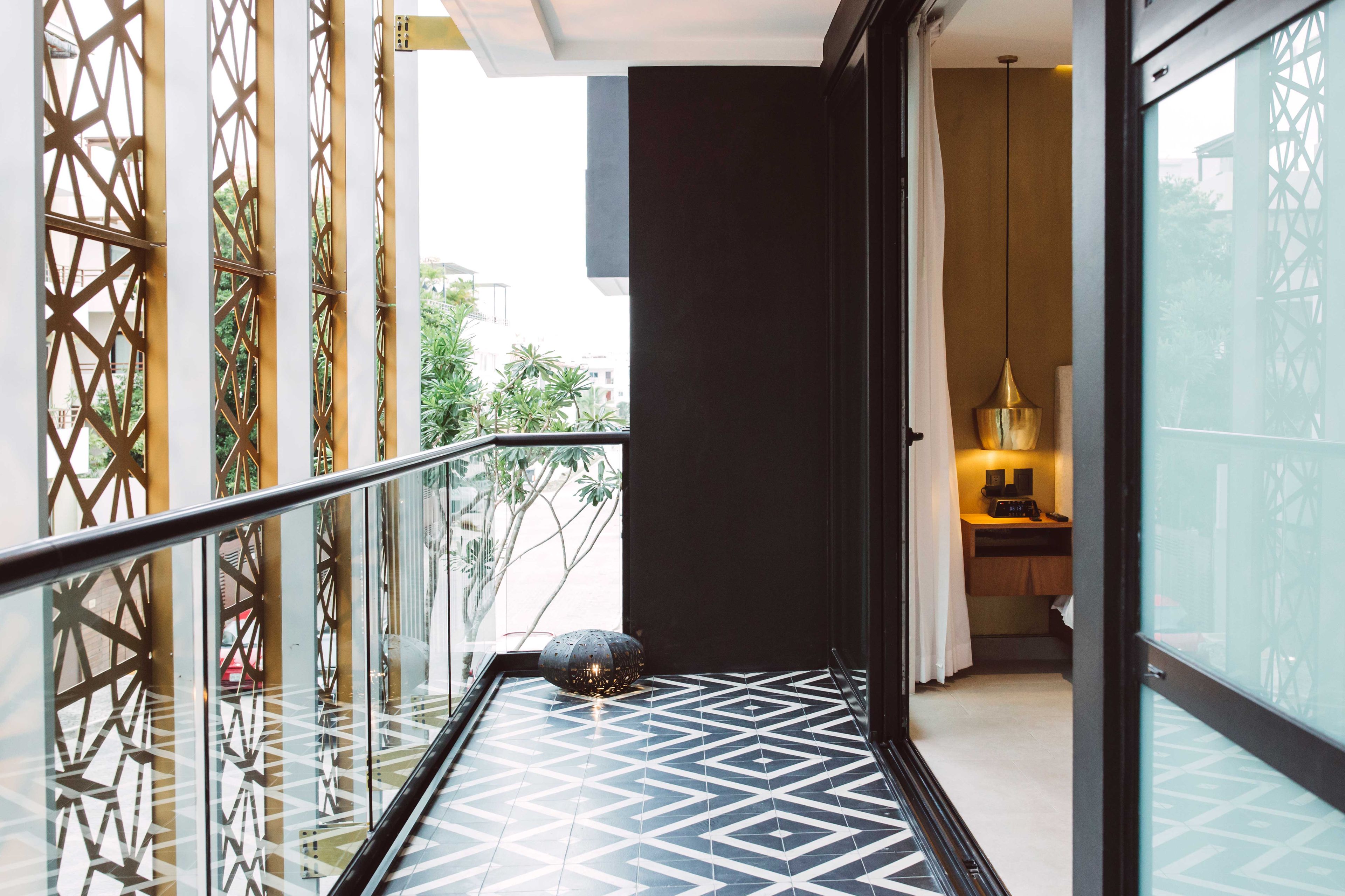 A modern hotel corridor with geometric patterned flooring, a glass door leading to a room with a bed and a nightstand, and a decorative lamp.