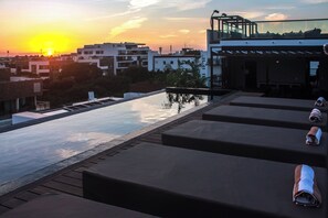 Un espace piscine aménagé sur le toit, avec des chaises longues et une balustrade en verre offrant une vue sur le paysage urbain au coucher du soleil.
