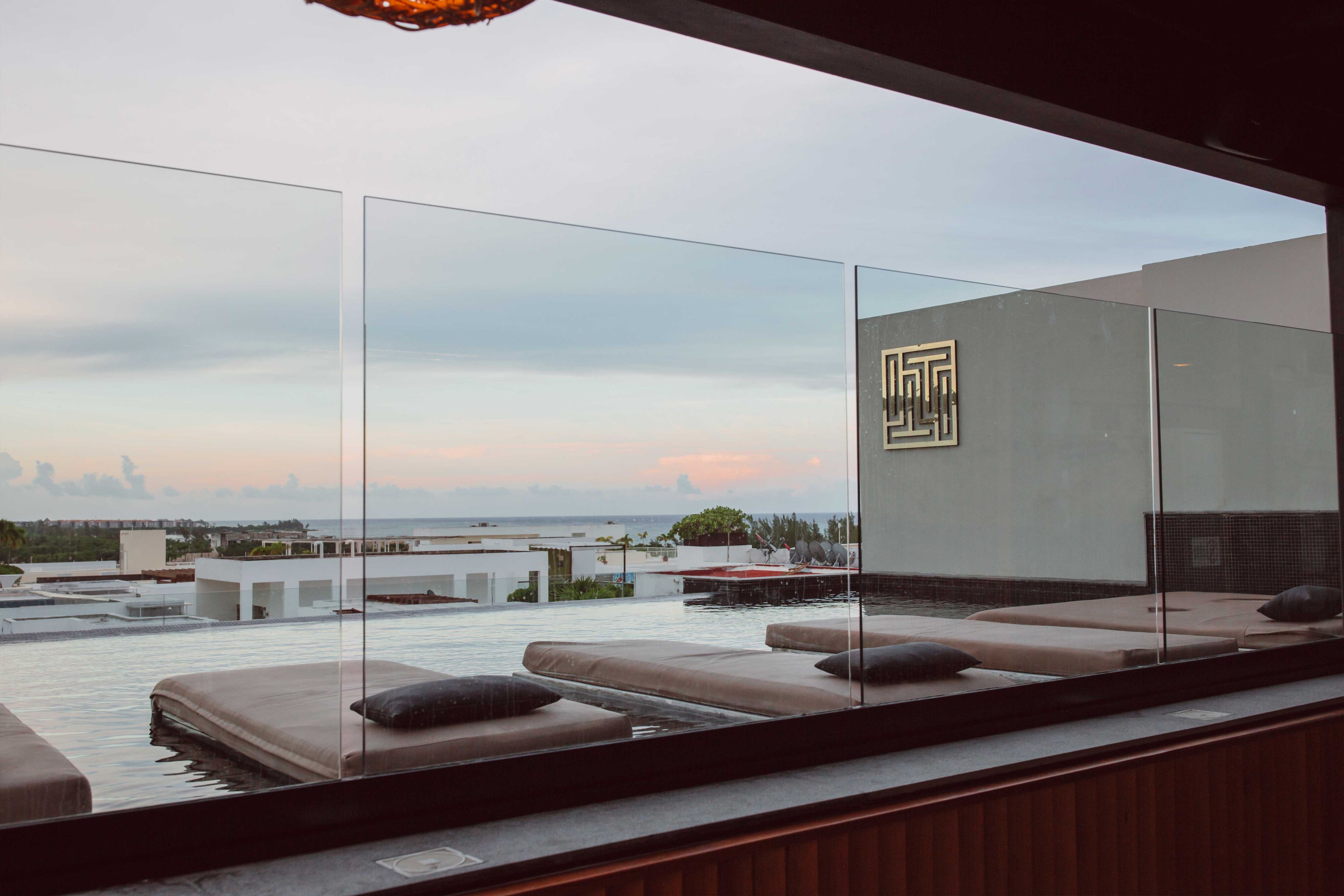 Un espace piscine moderne situé sur le toit, avec des murs en verre, un coin salon doté de coussins et une vue sur le paysage urbain.