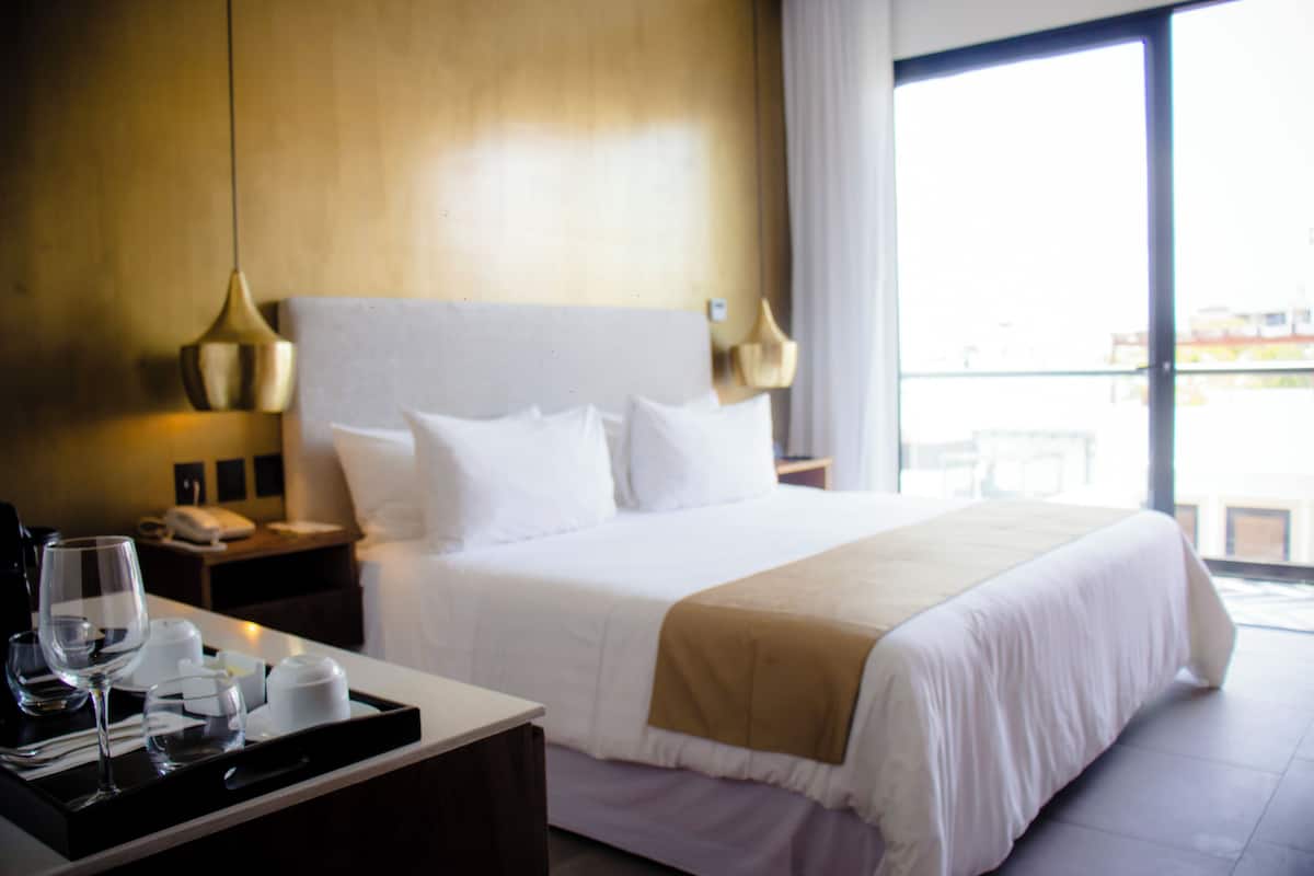 Habitación de hotel con una cama grande, una mesita de noche con lámpara, una bandeja con vasos y una taza, y vista al exterior a través de un amplio ventanal.