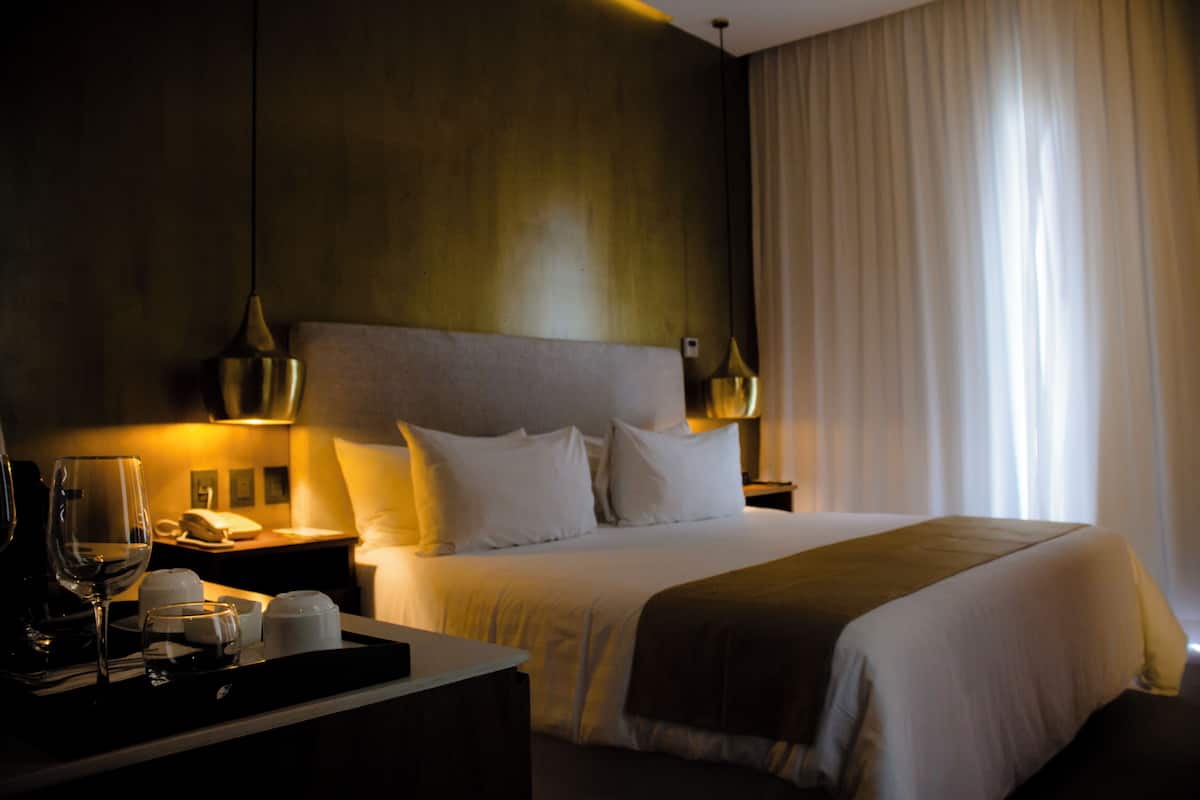 Una habitación de hotel con cama, mesitas de noche, una lámpara y un ventanal con cortinas.