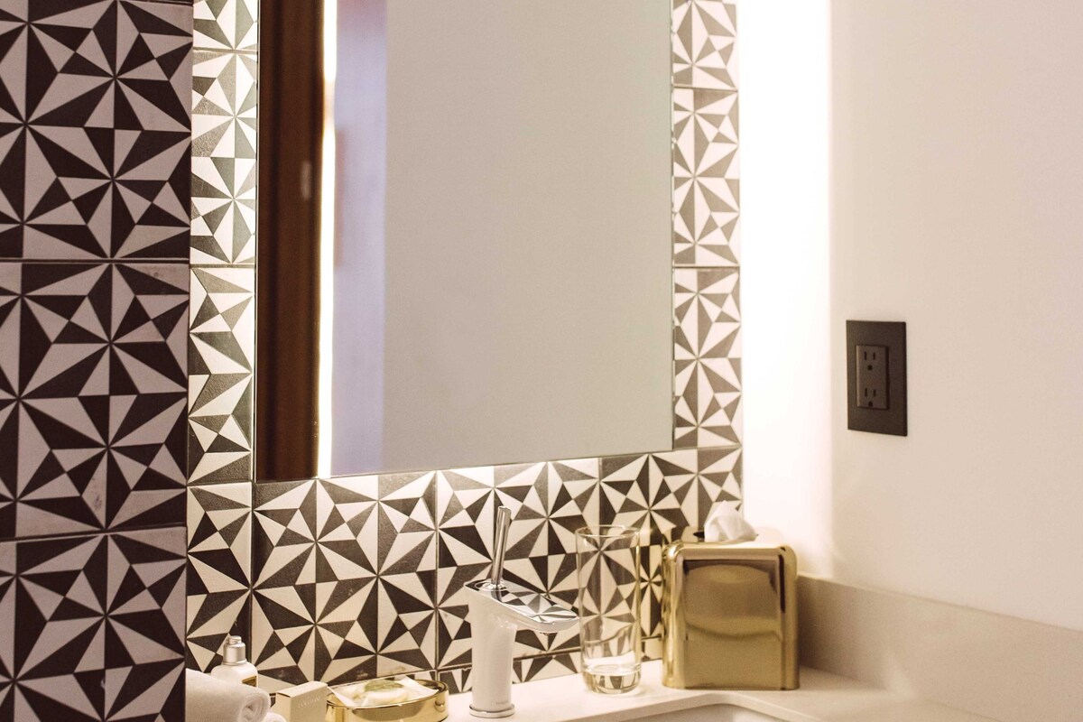 A bathroom with a geometric patterned wall, a large mirror, a white sink, and a gold toiletry bag.