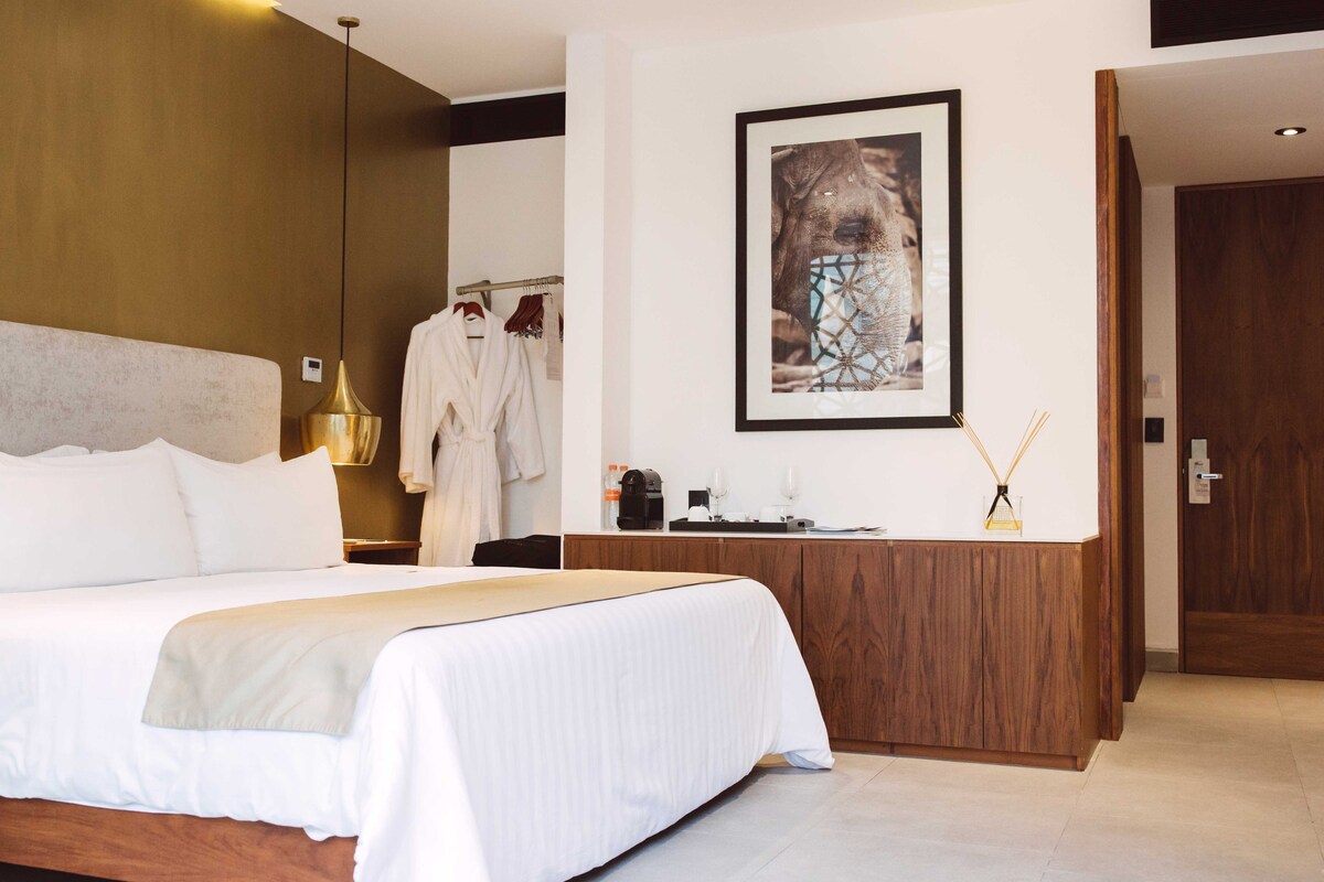 Habitación de hotel con una cama grande, cabecera de madera, un escritorio de madera, una obra de arte enmarcada en la pared y una bata colgada en un perchero.