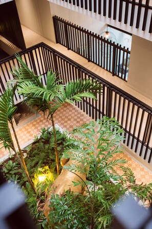 Un espacio interior de varios niveles con un atrio central que presenta un suelo con diseño, barandillas negras y abundante vegetación verde.