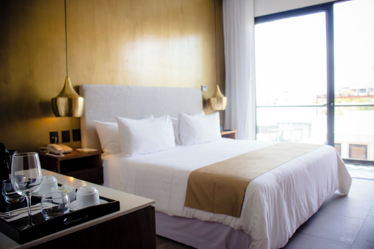 A hotel room with a large bed, a bedside table with a lamp, a tray with glasses and a cup, and a view of the outside through a large window.