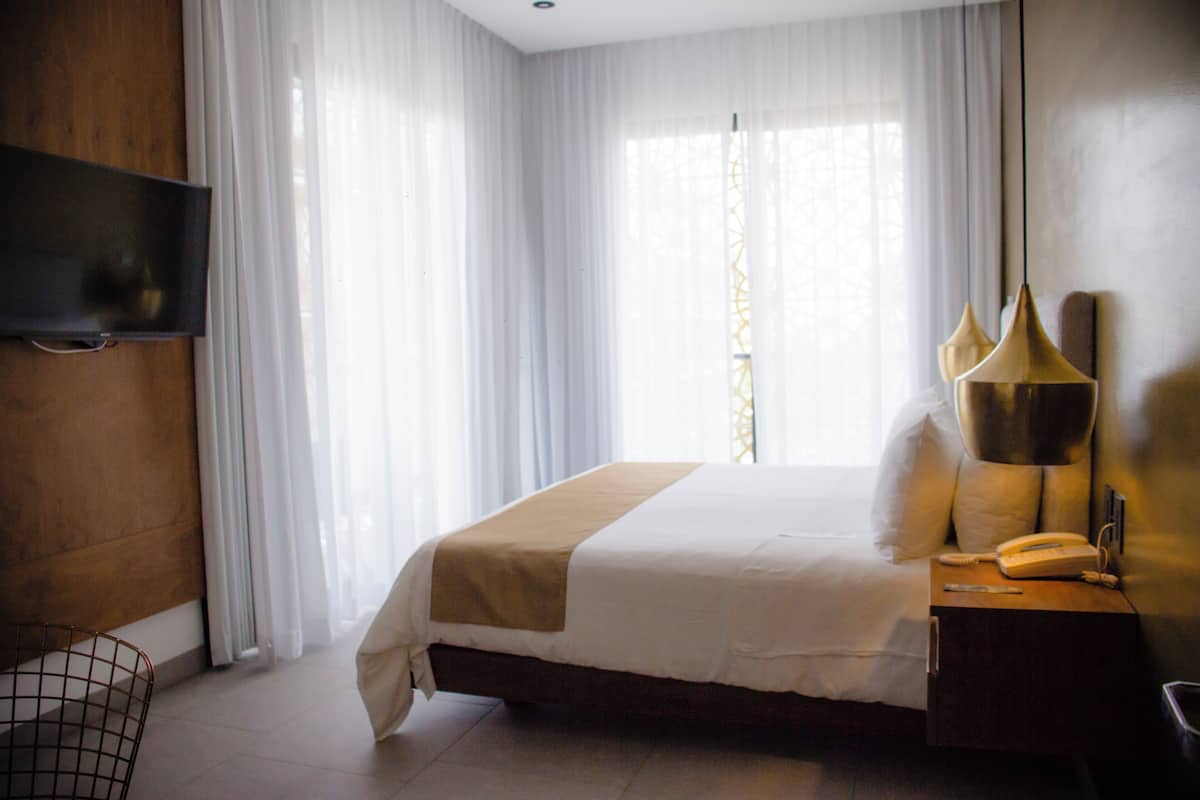 Habitación de hotel con una cama, mesita de noche con lámpara, televisor montado en la pared y ventana con cortinas traslúcidas.