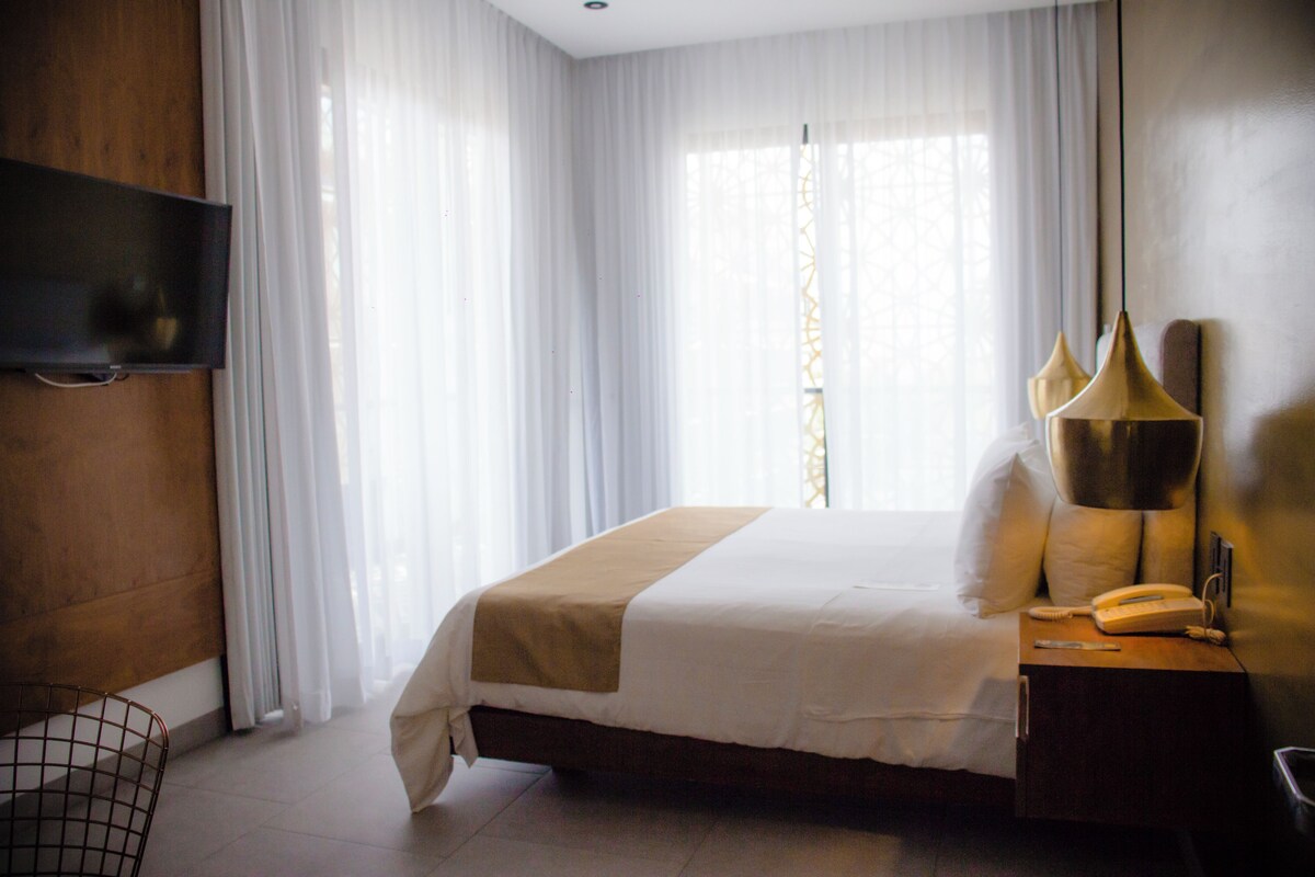 A hotel room with a bed, a bedside table with a lamp, a TV mounted on the wall, and sheer curtains.