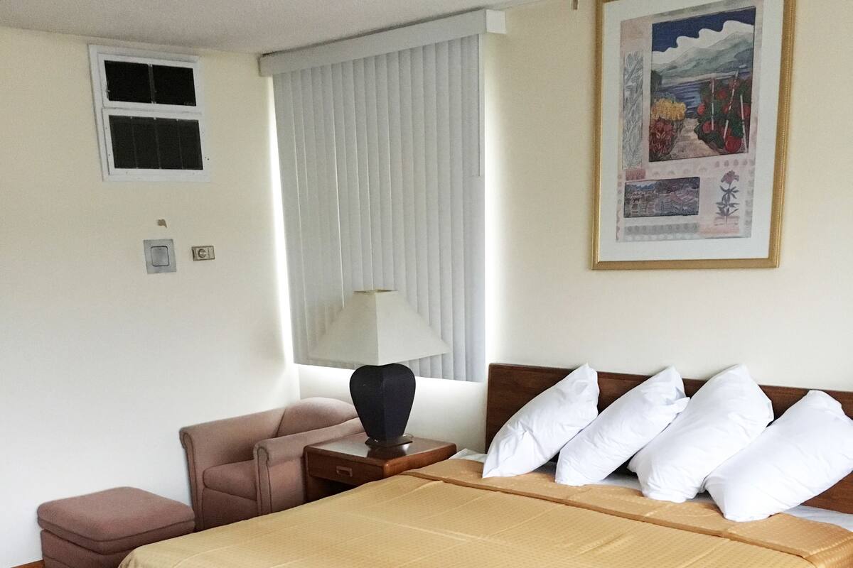 A hotel room with a large bed, a chair, a nightstand, and a window with blinds.