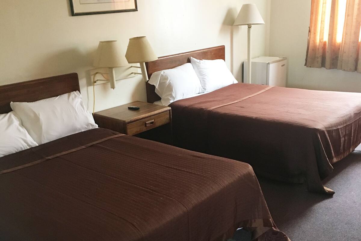 A hotel room with two single beds, a nightstand with a lamp, a small table, and a wall-mounted picture.