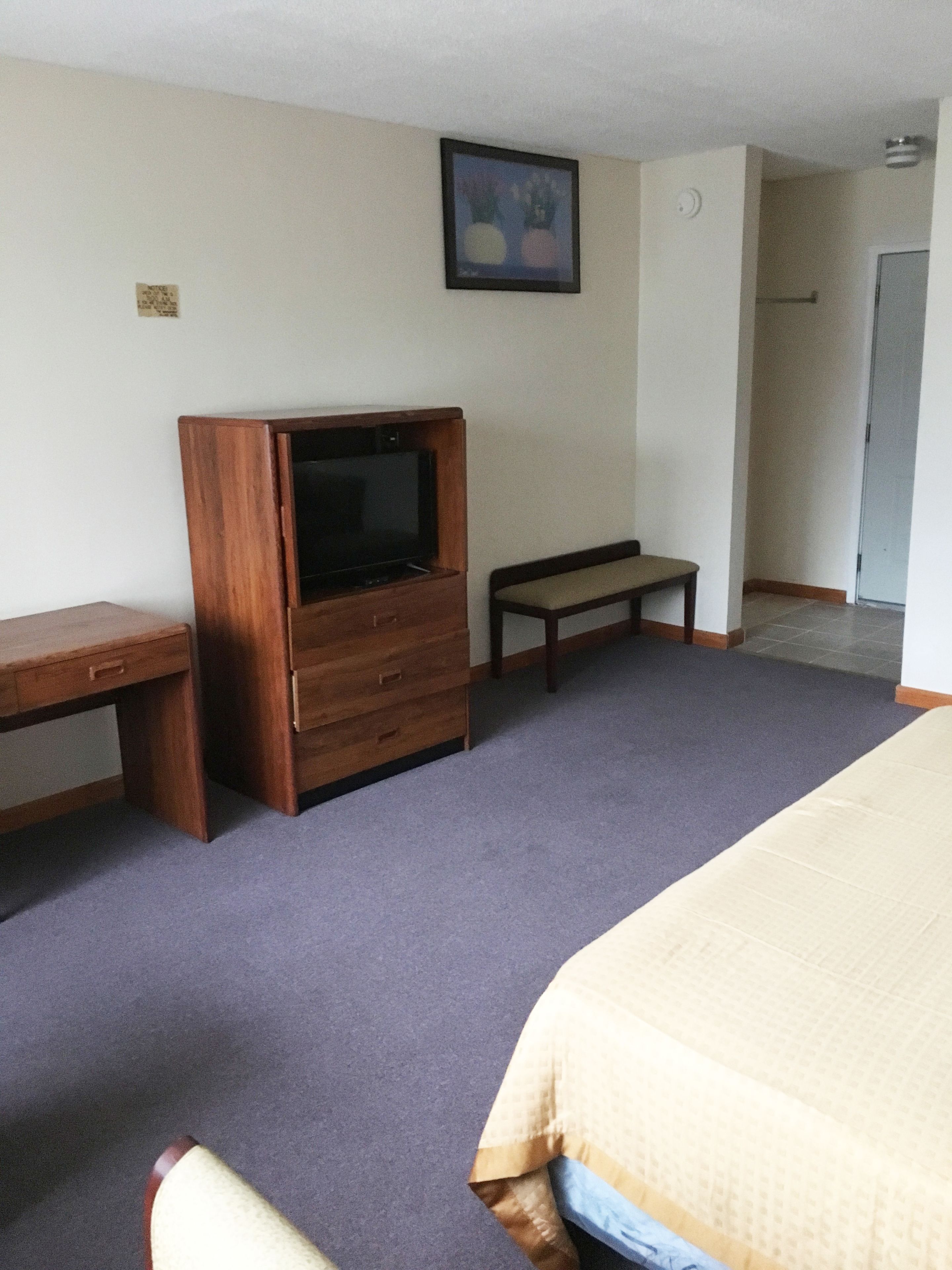 A hotel room with a bed, a wooden TV stand, a bench, and a desk.