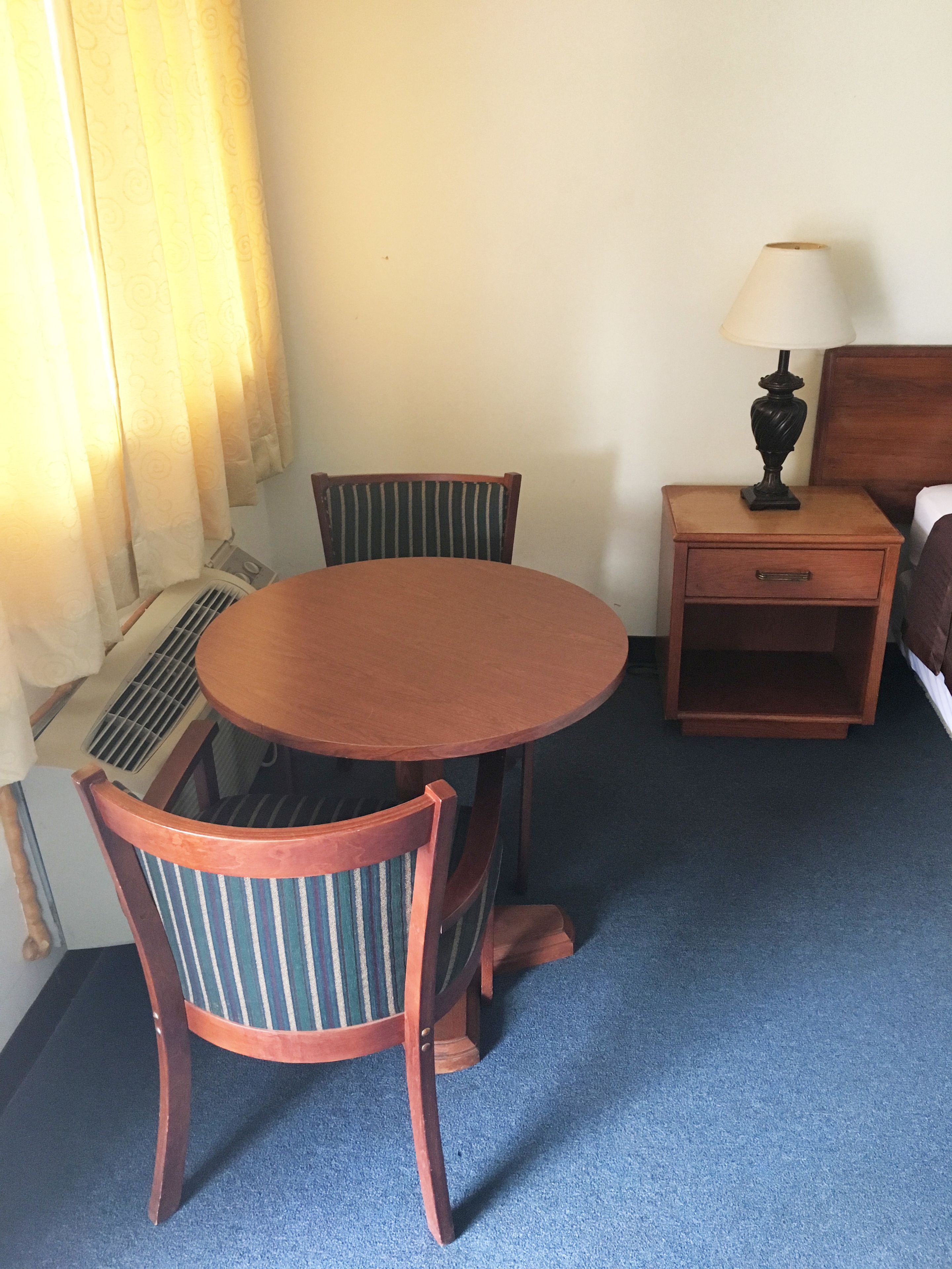 Ein Hotelzimmer mit einem runden Holztisch, zwei gestreiften Stühlen, einem Nachttisch mit Lampe und einem Bett mit Kopfteil.