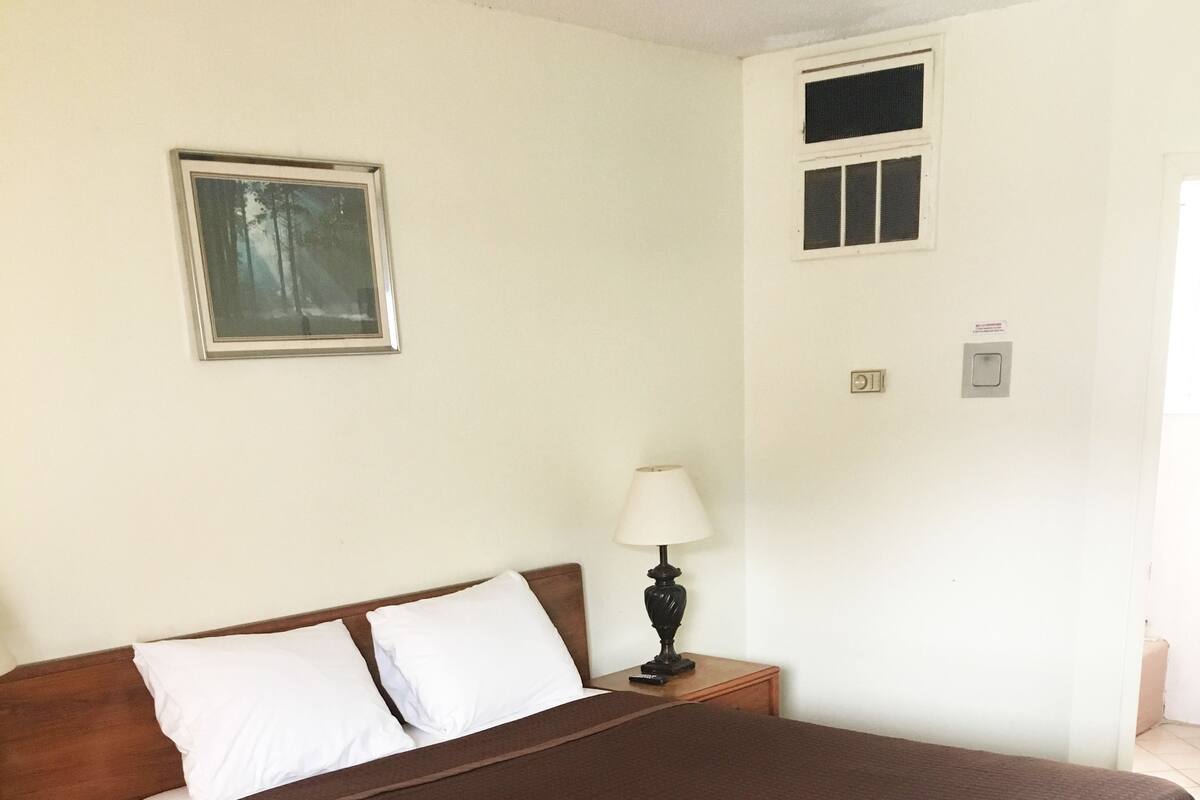 A single bed with a brown bedspread, a wooden nightstand with a lamp, a small table, and a framed picture on the wall.