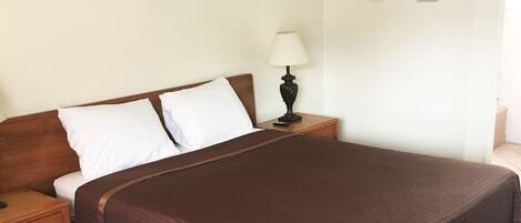 A single bed with a brown bedspread, a wooden nightstand with a lamp, a small table, and a framed picture on the wall.