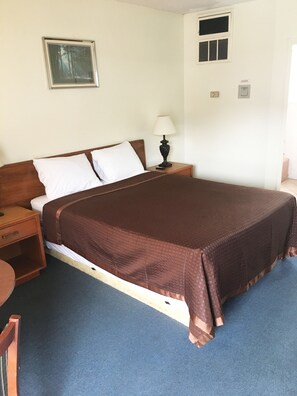 A single bed with a brown bedspread, a wooden nightstand with a lamp, a small table, and a framed picture on the wall. - Village Motel (Ellenville)