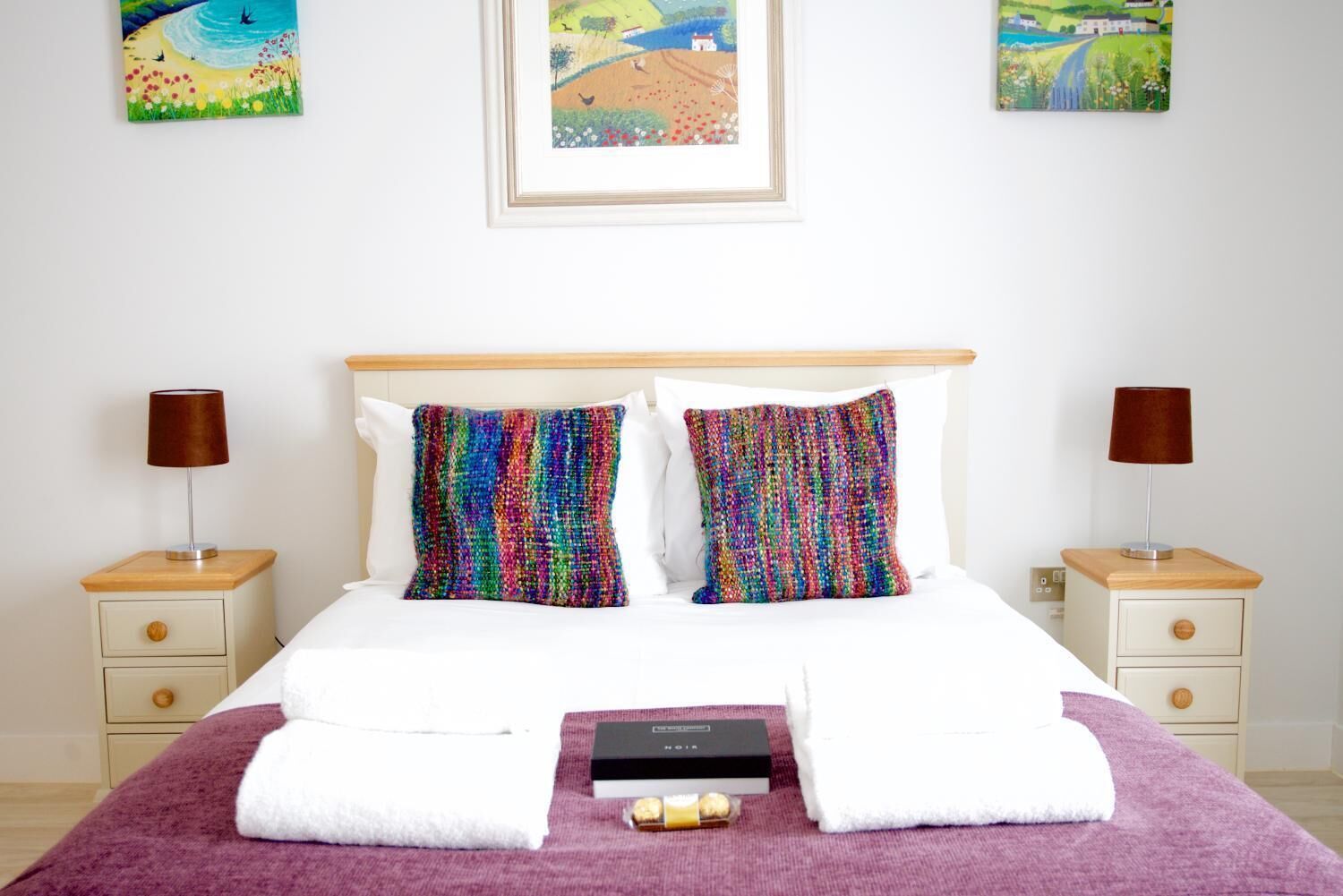 Una cama bien hecha con dos almohadas, un edredón morado y un juego de artículos de aseo sobre una mesita de noche de madera.