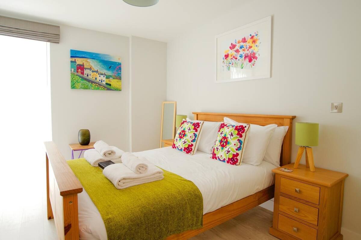 A neatly made bed with white linens and colorful pillows, a bedside table with a lamp, and a painting of a landscape on the wall.