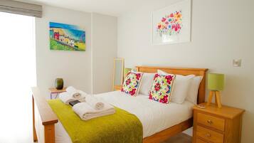 Una cama bien hecha con sábanas blancas y almohadas coloridas, una mesita de noche con lámpara, y un cuadro de un paisaje en la pared.