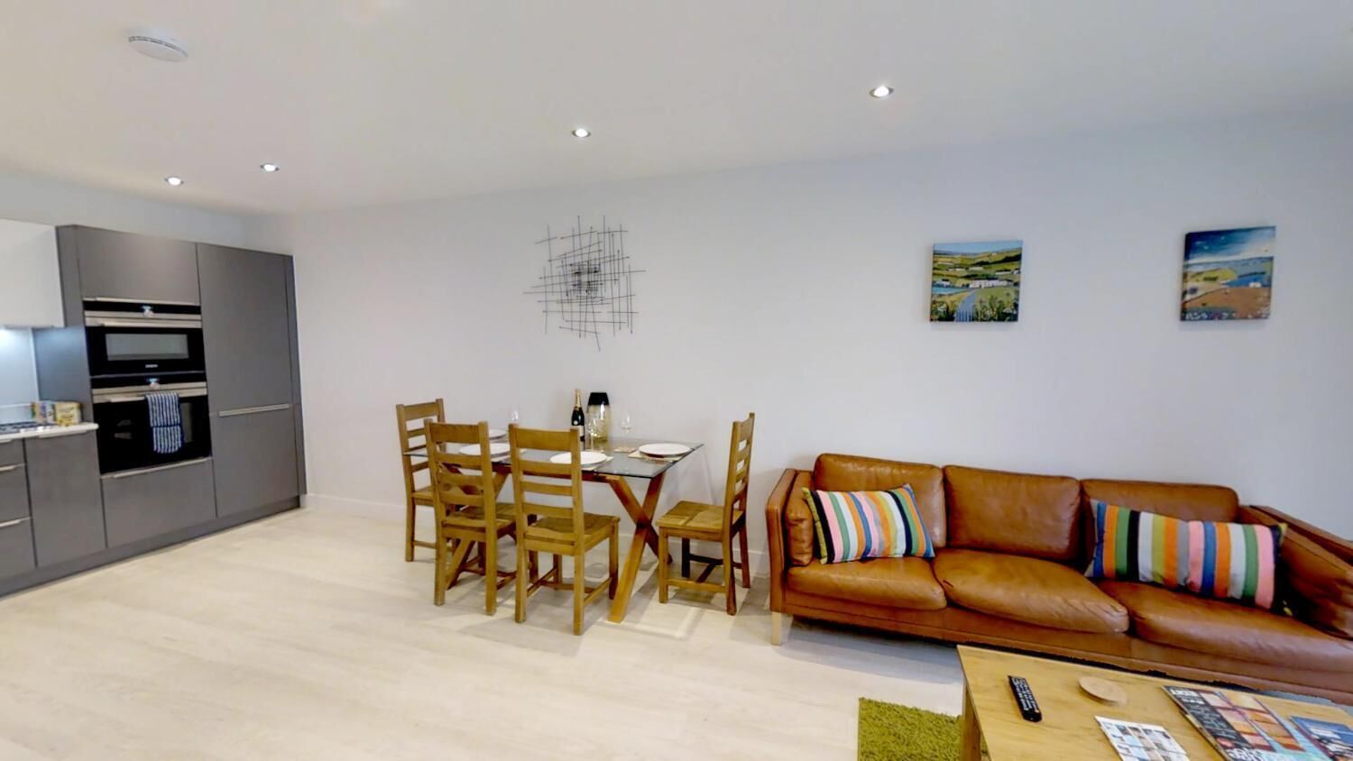 Una sala moderna con un sofá de cuero marrón, una mesa de comedor de madera y sillas. Hay una zona de cocina con armarios grises y un horno empotrado.